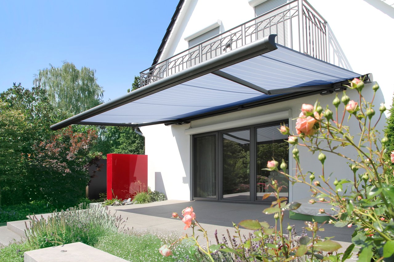 markilux 6000 mit blaugrauem Tuch beschattet die Terrasse eines weißen Einfamilienhauses mit Garten, Rosen und Lavendel.